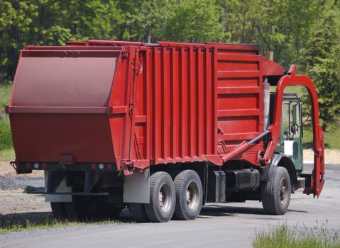 A large red refuse collection truck parked on a paved surface with a background of green trees and foliage, suggesting an outdoor setting in a rural or suburban area. The truck features a rectangular, metal waste container with a smooth top and ribbed sides, designed for rubbish collection and disposal. The container’s surface appears painted with a matte finish, showing slight texture from the metal panels. The truck has six wheels visible, with black tires and silver rims, and the wheel arches are also black. The cab, partially visible on the right side of the image, is white with a side mirror and window, indicating a standard commercial vehicle used by waste management services in Wimbledon or nearby areas. The environment appears well-lit by natural daylight, highlighting the vibrant red colour of the waste container, typical of rubbish removal vehicles used in the UK, including services like Commercial Waste Wimbledon, for efficient business waste collection across the town.