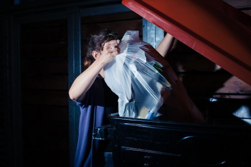 A young individual with short, dark hair is seen disposing of a large clear plastic bag filled with various waste items into a black industrial rubbish container. The container is situated outdoors on a paved surface, with a background of wooden fencing and a dark metal structure, indicating a typical waste disposal area in Wimbledon. The plastic bag contains crumpled papers and miscellaneous debris, and the person appears to be lifting the lid of the container to deposit the waste. The scene is illuminated by natural light, casting soft shadows and highlighting the textures of the plastic, the wooden fencing, and the metallic container. This image exemplifies waste management activities that a rubbish removal service like Commercial Waste Wimbledon handles, particularly in a busy town setting, demonstrating careful disposal practices and associated environmental considerations.