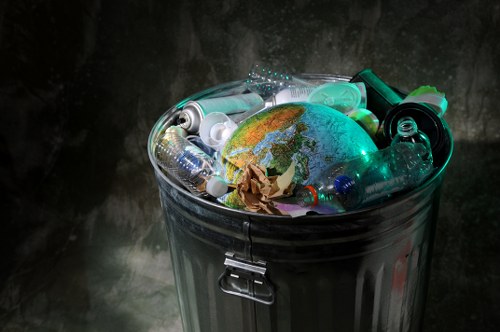 A close-up view of an open metal rubbish bin placed outdoors on a dark, slightly textured surface. The bin is filled with various discarded plastic bottles, containers, and a crumpled brown paper bag. The plastic waste includes transparent bottles, some with caps, and different shapes and sizes, with textures ranging from smooth to ridged. A small, plastic globe model depicting Earth is situated among the debris, partially obscured by the bottles. The scene suggests the collection of recyclable waste, highlighting typical items found in rubbish removal services in Wimbledon or similar urban areas. The lighting creates subtle reflections on the metallic surface of the bin and the shiny plastics, emphasizing the different materials. This image visually represents waste collection and disposal activities associated with commercial rubbish management in a metropolitan environment, with the backdrop being a dark wall or surface that contrasts with the clutter of refuse.