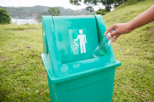 A turquoise recycling bin with a white icon of a person disposing of waste, situated outdoors on a grassy area near a scenic body of water and green hills. A hand holds a clear plastic bottle, about to place it into the bin. The environment features natural light, with the background showing lush trees and a distant shoreline, indicating a park or riverside location. The bin appears to be designed for recyclable waste, aligning with waste management and rubbish removal services provided by companies such as Commercial Waste Wimbledon.