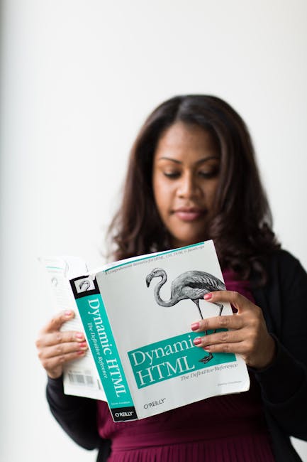 A woman with long dark hair and medium skin tone is holding a paperback book titled 'Dynamic HTML' by O'Reilly, featuring a detailed illustration of a flamingo on the cover. She is standing indoors ag