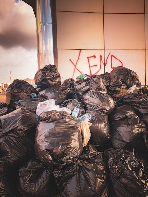 A large pile of black refuse bags, some torn open, stacked against a tiled wall in an outdoor urban setting. The bags appear full and are made of glossy plastic, with some containing visible waste suc