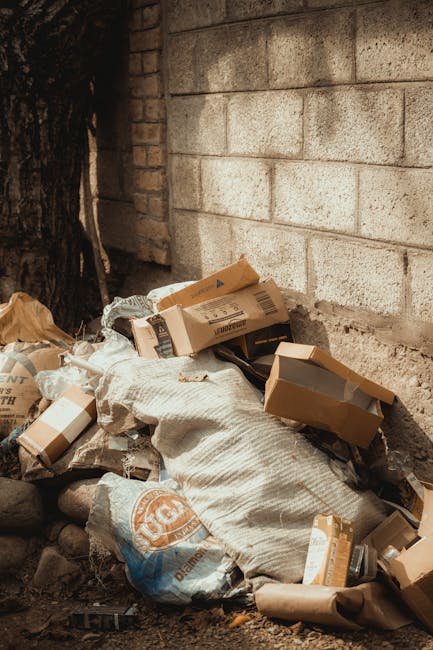 A large collection of mixed waste and rubbish, including flattened cardboard boxes, plastic bags filled with waste, paper and packaging materials, and larger items such as a blue plastic storage conta