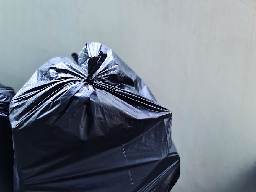 Close-up image of a black plastic rubbish bag, tightly knotted at the top, resting against a plain, light-colored wall. The bag appears to be made of shiny, durable plastic with visible creases and sl